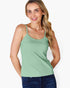 A woman wearing a light sage green spaghetti-strap tank top and dark blue jeans, standing against a plain white studio background.