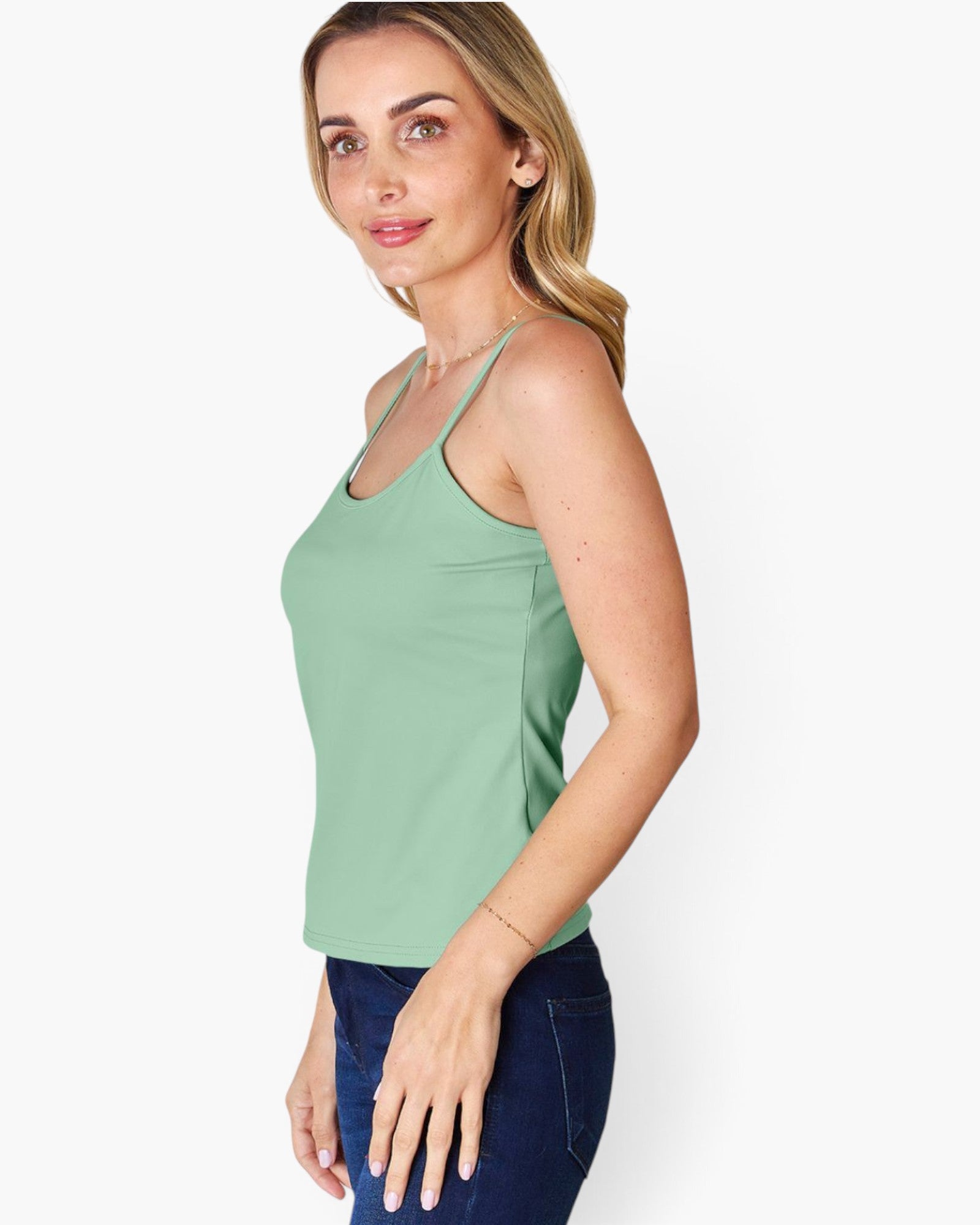 A woman wearing a sage green sleeveless tank top and dark blue jeans, shown from a side profile against a light gray studio background.