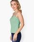 A woman wearing a sage green sleeveless tank top and dark blue jeans, shown from a side profile against a light gray studio background.