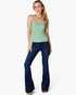 A woman modeling a light sage green spaghetti-strap tank top and dark blue flare jeans against a plain white studio background.