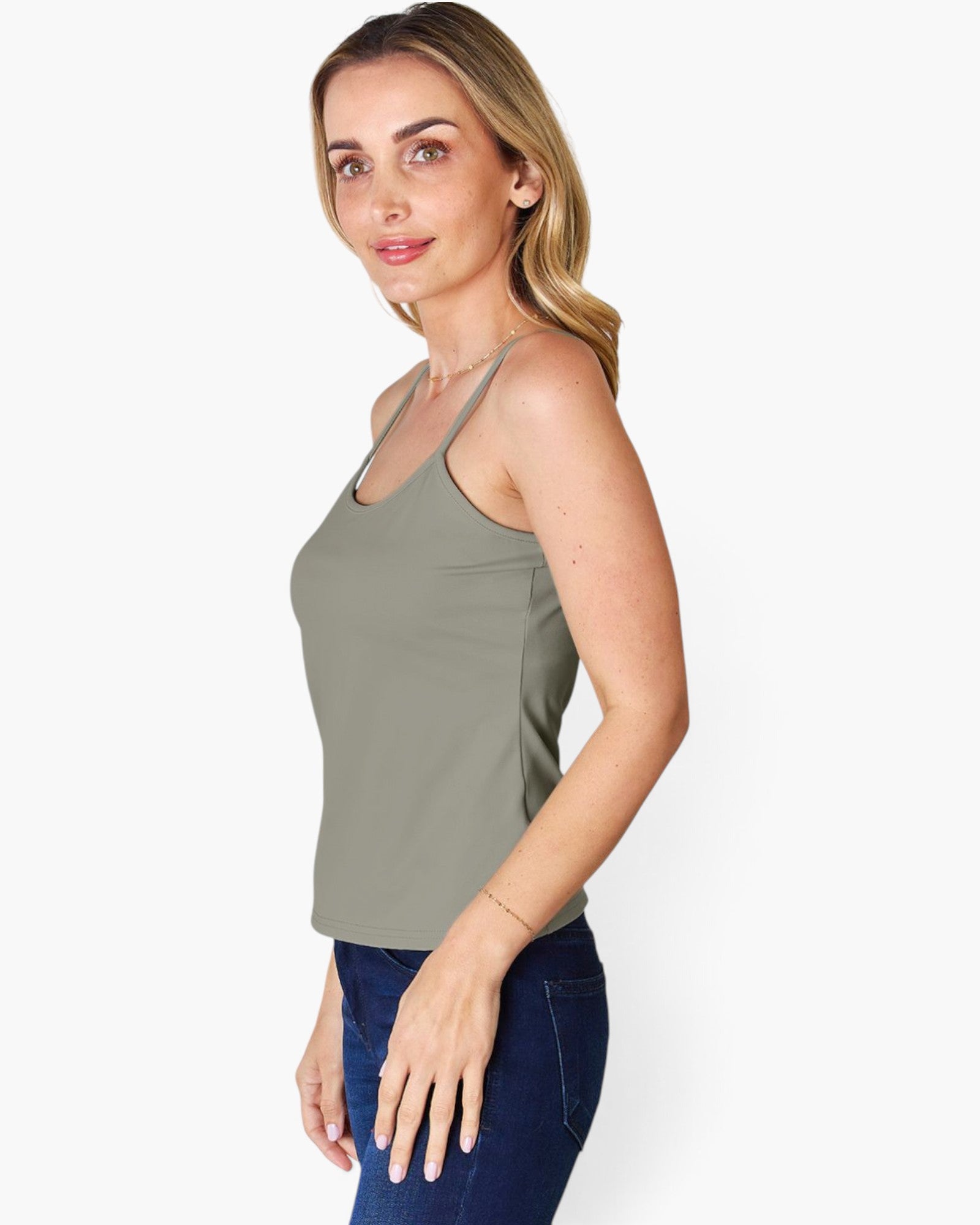 A woman modeling a sleeveless scoop-neck tank top in a muted sage green color, paired with dark blue jeans, against a plain white studio background.