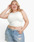 A plus-size blonde woman wearing a fitted white sleeveless crop top and light blue distressed denim shorts, posing against a light gray studio background.