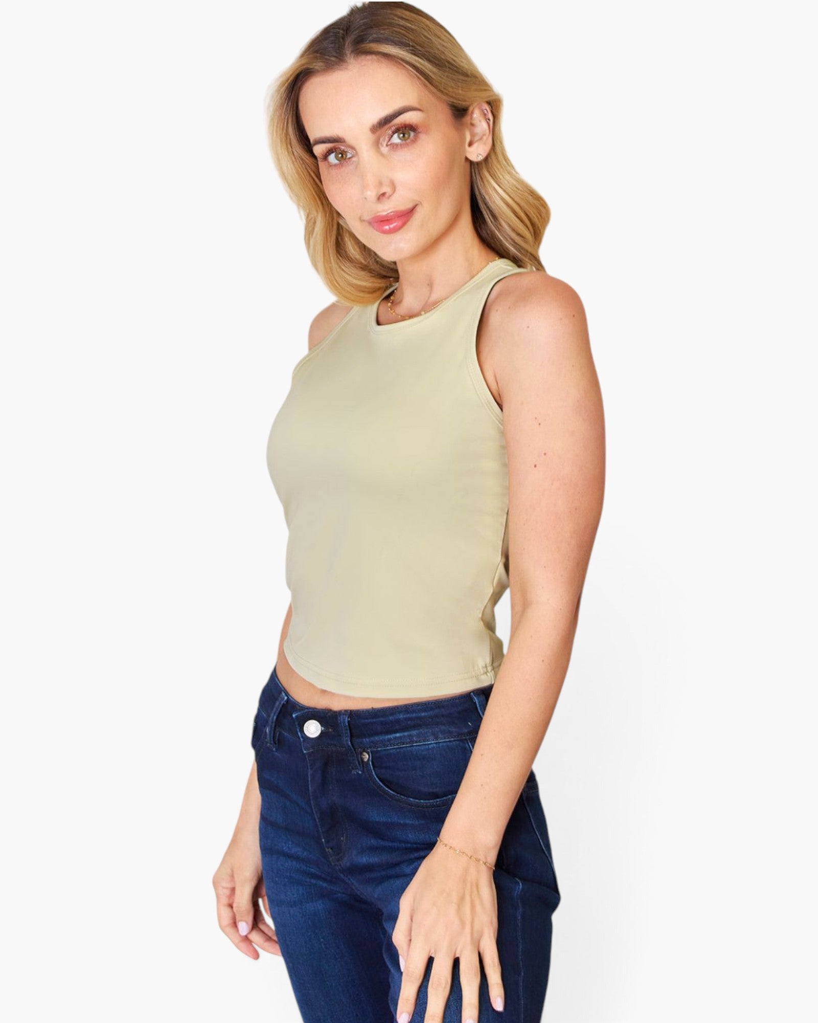 A woman modeling a sleeveless, cropped knit top in a light sage green color, paired with dark blue jeans, against a plain white studio background.
