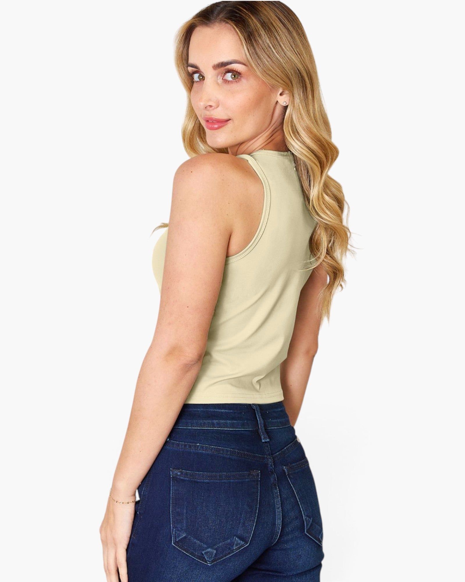 A woman with blonde wavy hair is shown from the back wearing a light beige sleeveless tank top and dark blue jeans against a plain white background.