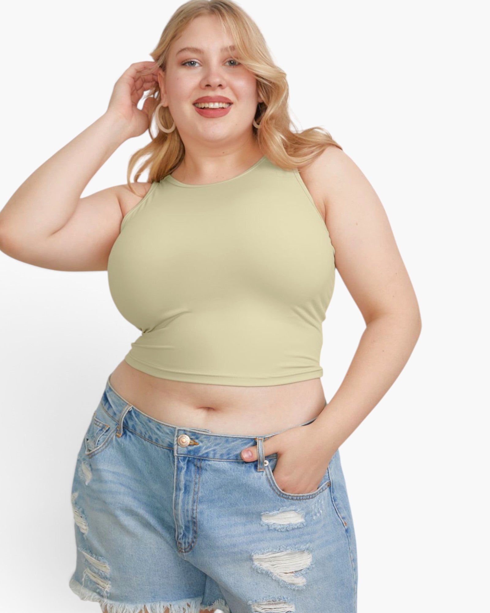 A plus-size blonde woman wearing a pale sage green sleeveless crop top and light-wash distressed denim shorts, posing against a solid white studio background.