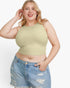 A plus-size blonde woman wearing a pale sage green sleeveless crop top and light-wash distressed denim shorts, posing against a solid white studio background.