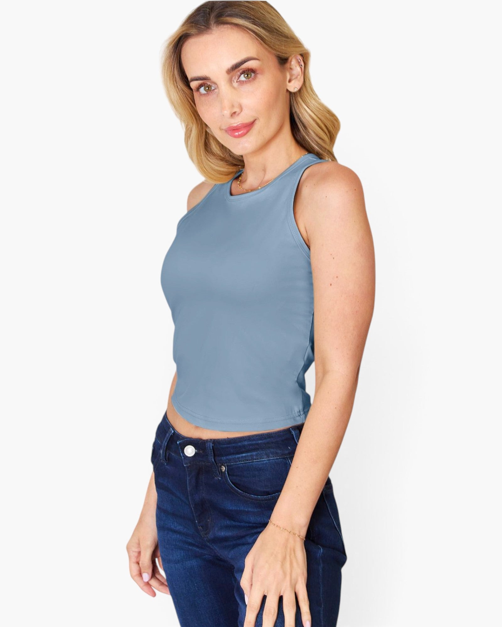 A woman modeling a sleeveless, scoop-neck crop top in a dusty slate blue color, paired with dark blue jeans, against a plain white studio background.