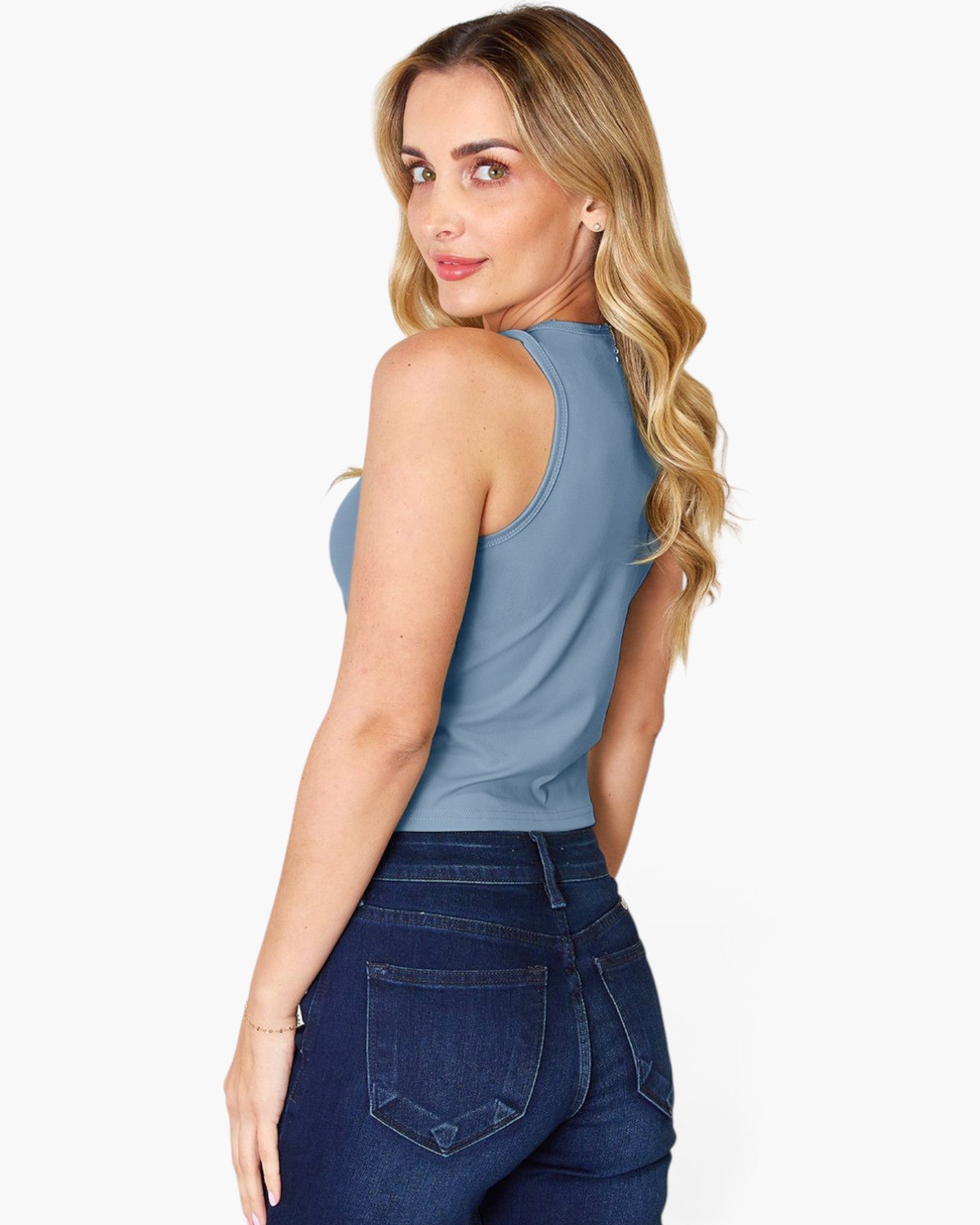 A woman with blonde wavy hair is shown from the back wearing a dusty blue sleeveless tank top and dark blue jeans against a light gray background.