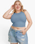 A plus-size woman wearing a dusty blue sleeveless crop top and light-wash distressed denim shorts, posing against a solid white studio background.