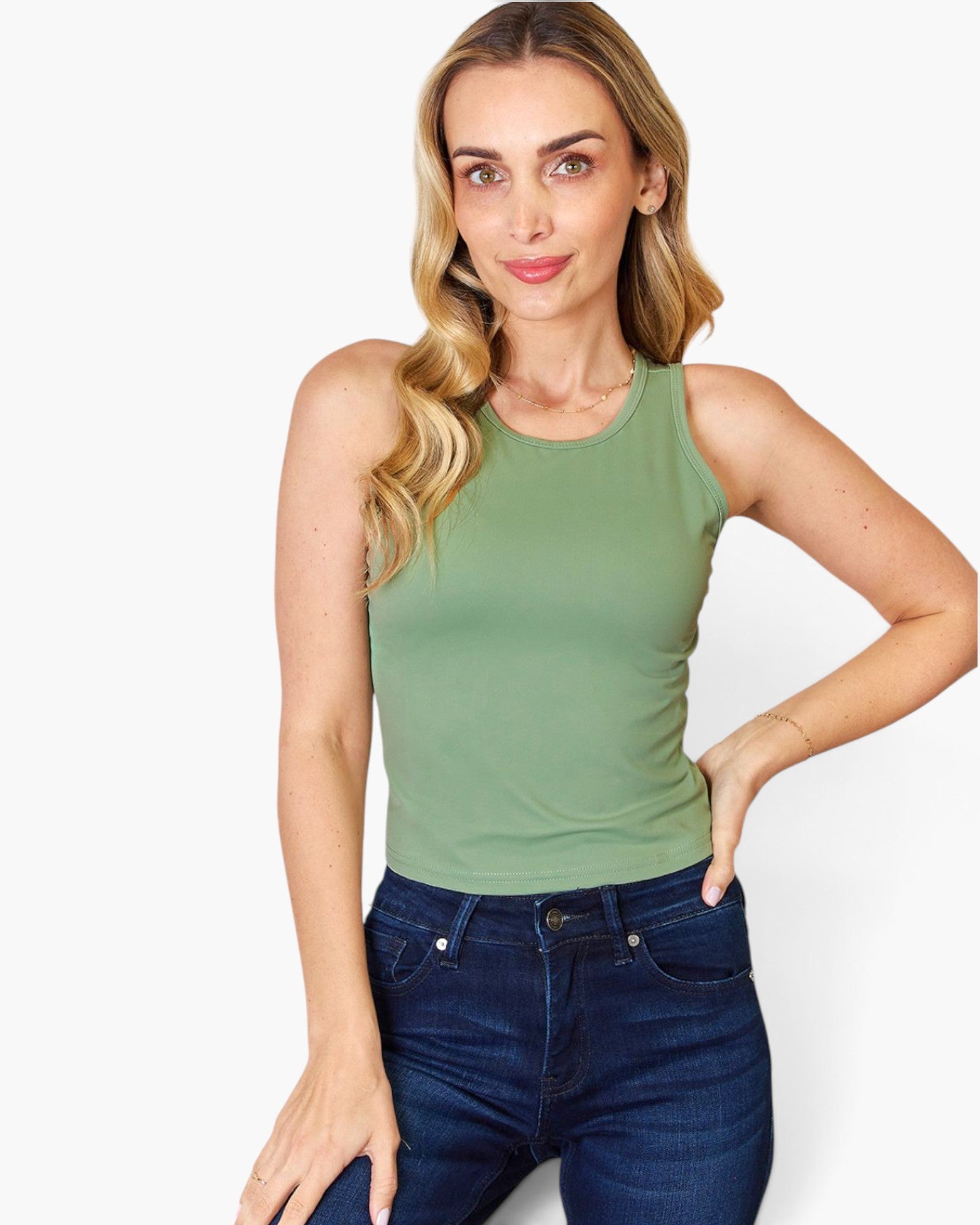 A woman modeling a sleeveless crew-neck tank top in sage green, paired with dark blue jeans, against a plain white studio background.