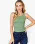 A woman modeling a sleeveless crew-neck tank top in sage green, paired with dark blue jeans, against a plain white studio background.