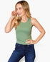A woman wearing a sage green sleeveless tank top and dark blue jeans, posing against a light gray studio background.
