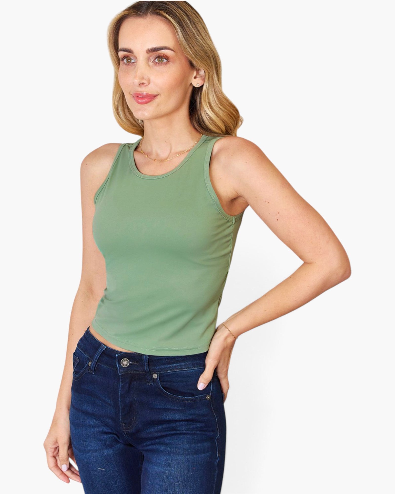 A woman modeling a sleeveless crew-neck tank top in sage green, paired with dark blue jeans, against a plain light gray background.