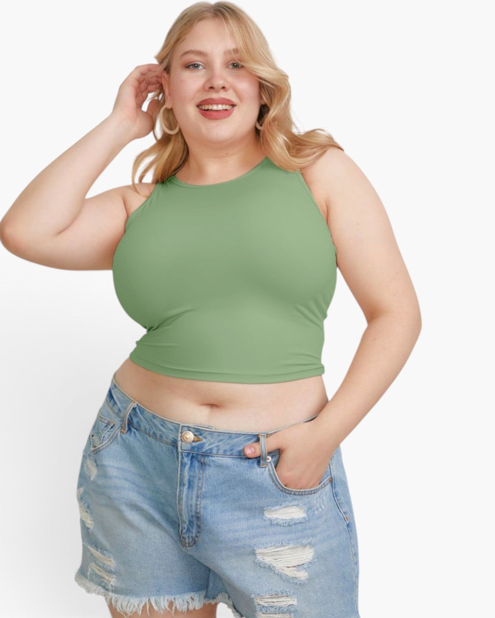 A plus-size blonde woman wearing a sage green ribbed halter crop top and light-wash distressed denim shorts, posing against a solid white studio background.