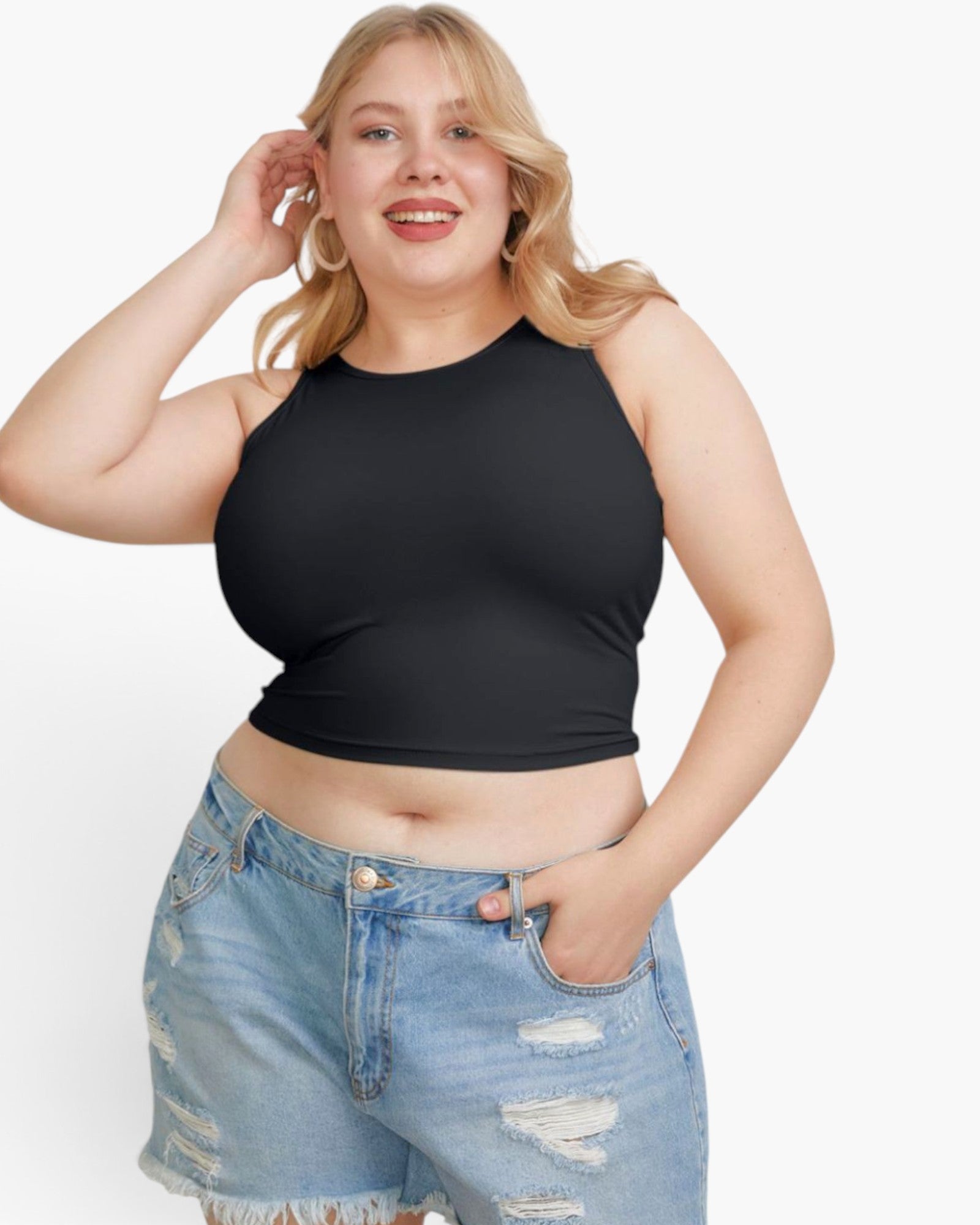 A plus-size blonde woman wearing a black sleeveless crop top and light-wash distressed denim shorts, posing against a solid white studio background.