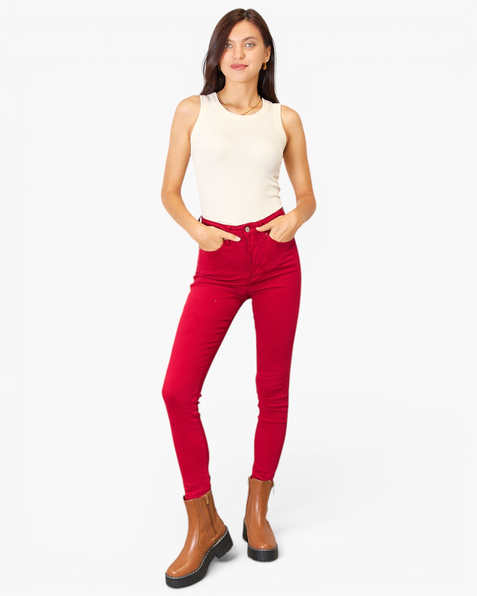 A woman modeling bright red skinny jeans with a cream sleeveless top and brown ankle boots, standing against a plain white studio background.