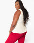 A woman with long, dark, curly hair is shown from a side-back angle, wearing a sleeveless cream-colored top and bright red pants against a plain white studio background.
