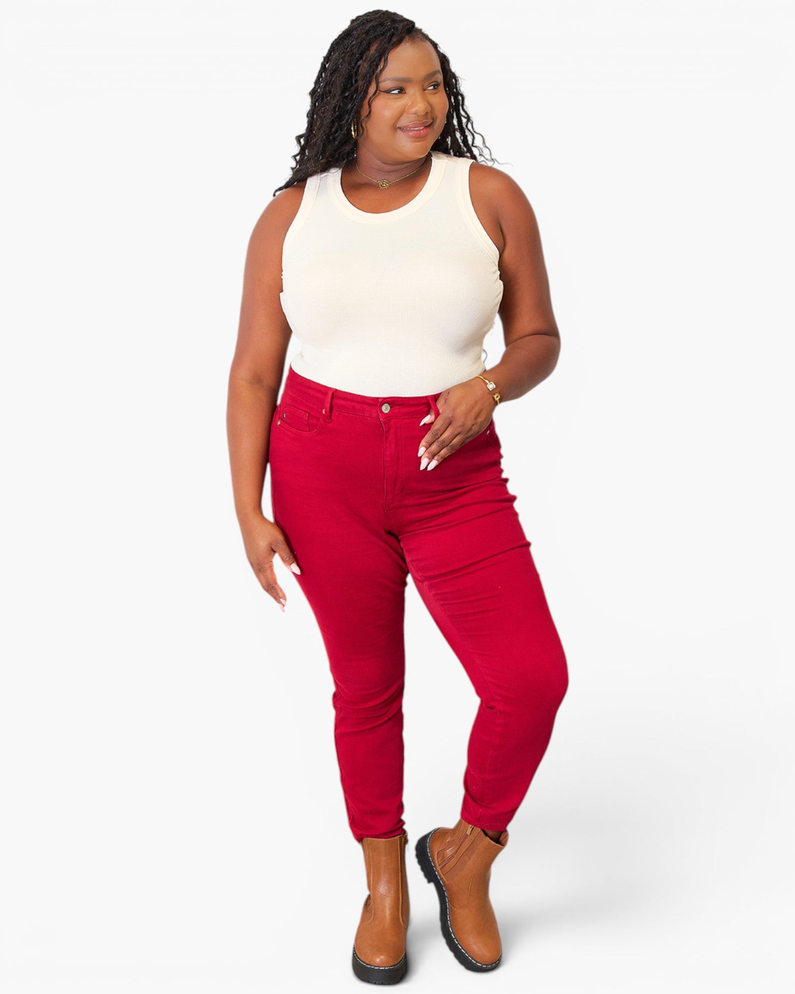 A woman modeling bright red skinny jeans with a white sleeveless top and brown ankle boots, standing against a light gray studio background.