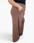 A person wearing loose, wide-leg brown pants with a smocked elastic waistband and side pockets, shown from the waist down against a light gray studio background.