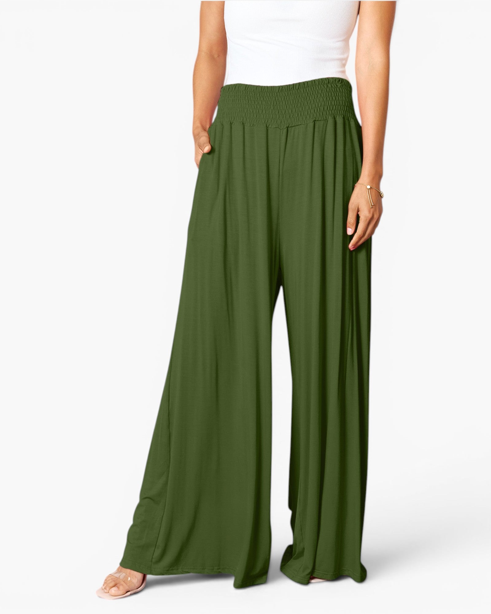 A model wearing a pair of wide-leg, high-waisted olive green pants with a smocked elastic waistband, styled with a white tank top and clear high-heel sandals against a light gray studio background.