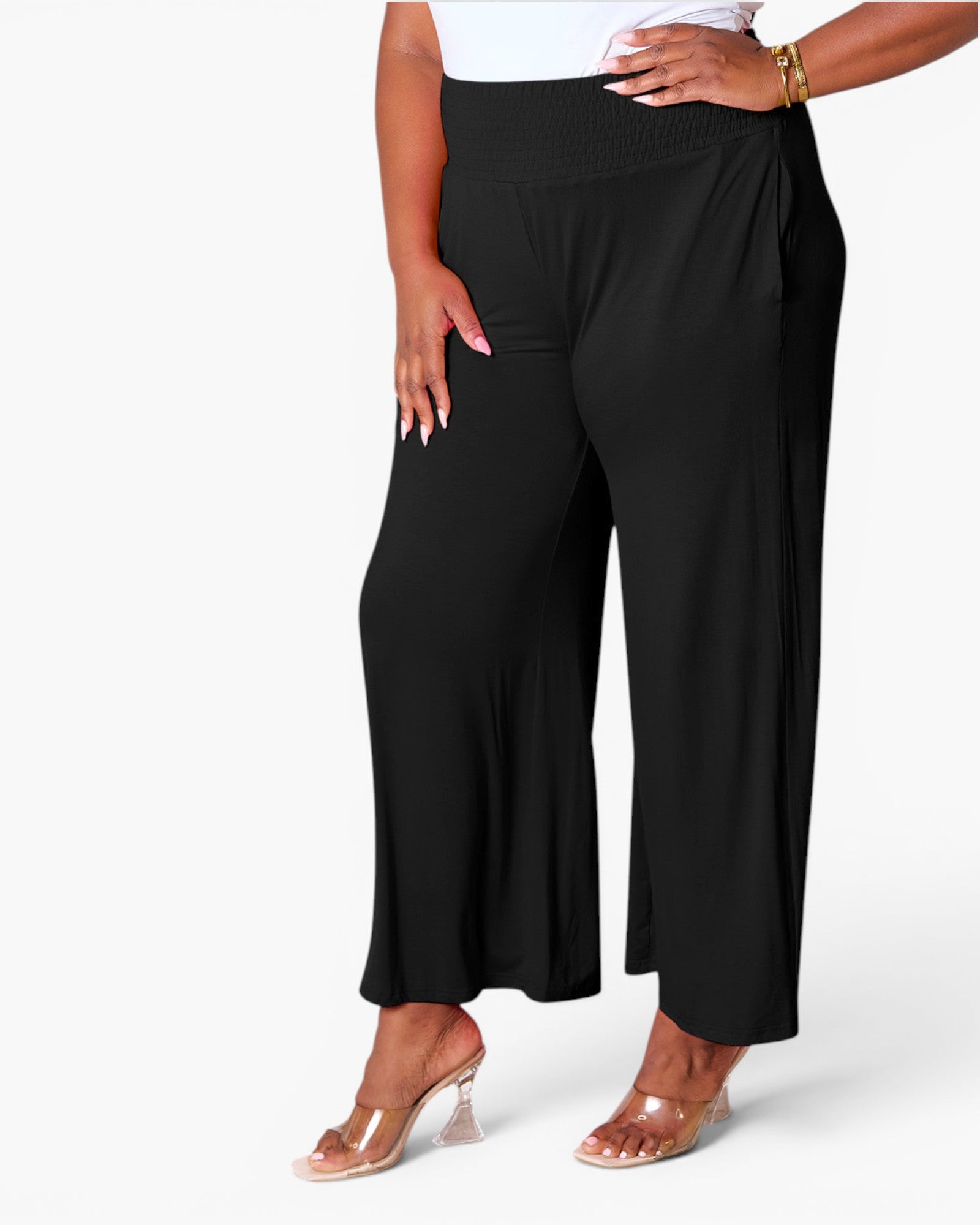 A model wearing black wide-leg cropped pants with a smocked elastic waistband, paired with a white top and clear high-heeled sandals, shown against a plain white studio background.