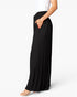 A side-profile view of a model wearing black wide-leg pleated maxi pants with a side pocket, paired with a white top and clear high-heeled sandals, against a plain white studio background.