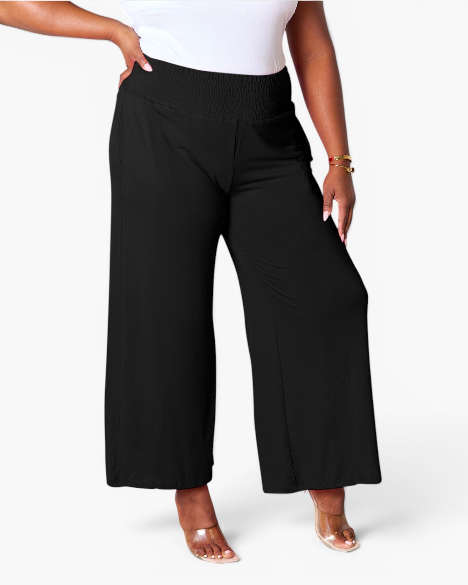 A model wearing black wide-leg cropped pants with a smocked elastic waistband, styled with a white top and clear high-heeled sandals against a light gray studio background.