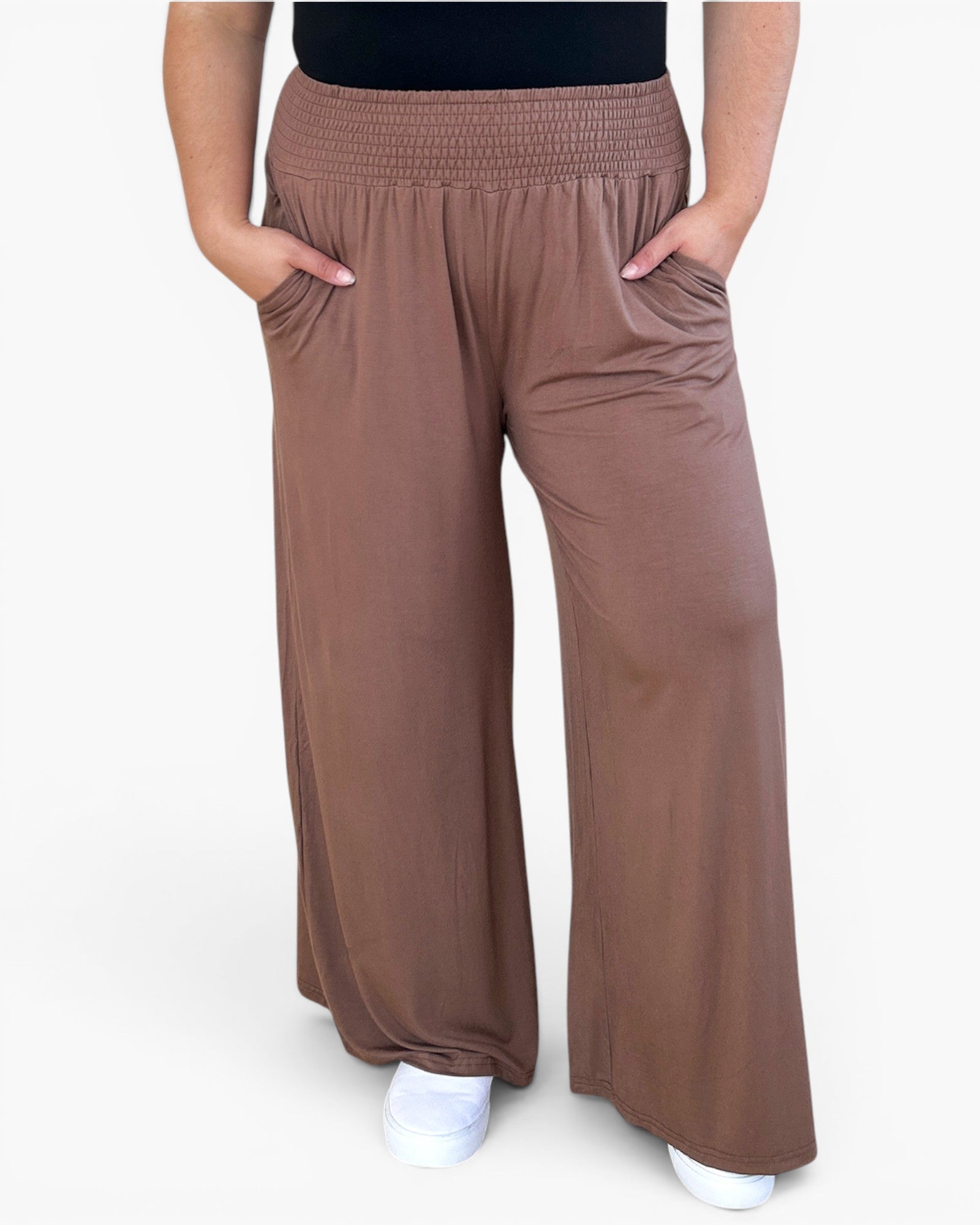 A person wearing loose, wide-leg brown pants with a smocked elastic waistband and side pockets, paired with a black top and white sneakers, against a plain light background.