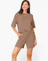 A woman modeling a matching lounge set consisting of a short-sleeve crewneck top and coordinating shorts in a solid taupe-brown color, set against a plain white studio background.