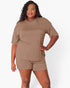 A woman modeling a matching two-piece lounge set consisting of a short-sleeve crewneck top and matching shorts in a solid taupe-brown color, set against a plain light gray studio background.