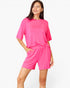 A woman modeling a bright pink lounge set consisting of a short-sleeve crewneck top and matching shorts against a plain white studio background.