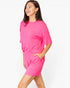 A woman modeling a bright pink lounge set consisting of a short-sleeve top with a chest pocket and matching shorts, against a plain white studio background.