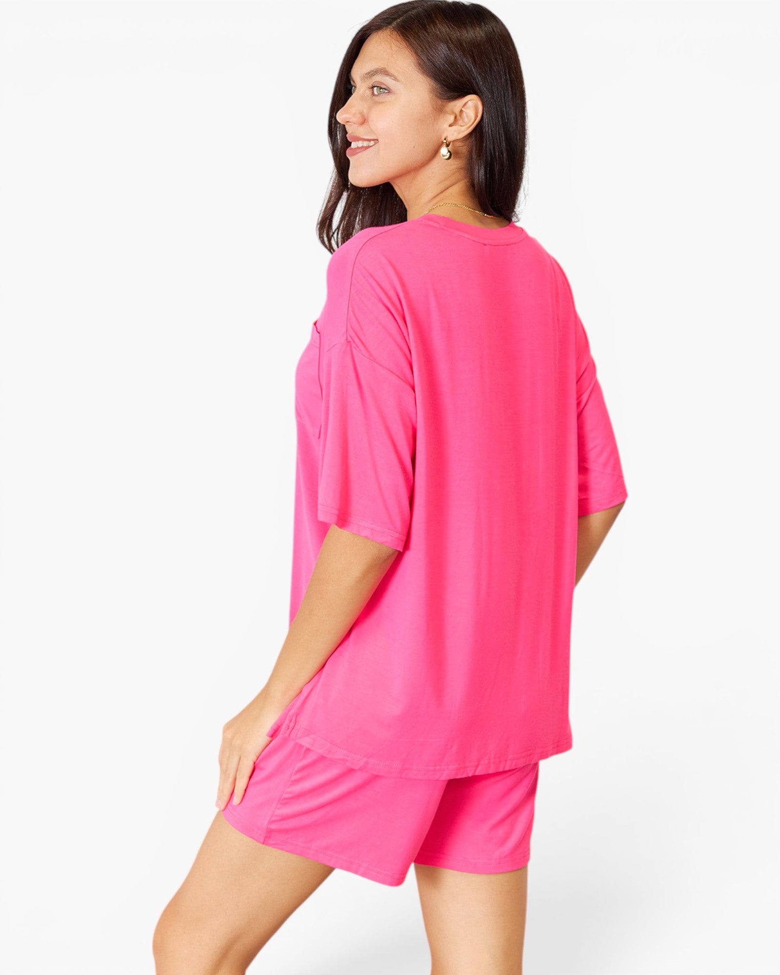 A woman modeling a bright pink lounge set consisting of an oversized short-sleeve top and matching shorts, shown from a three-quarters back view against a plain white studio background.