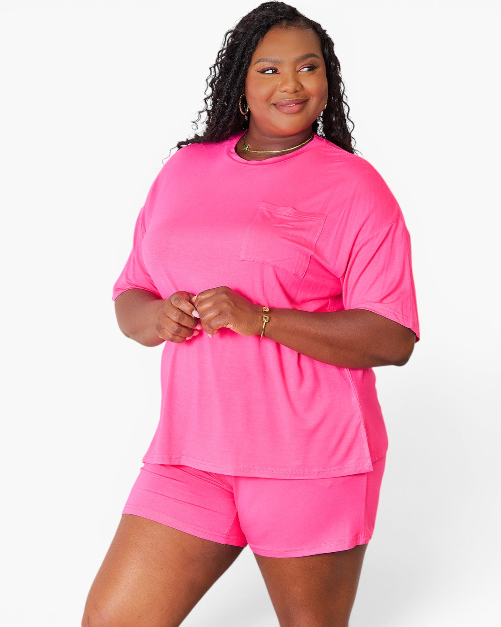 A woman modeling a bright pink matching lounge set consisting of a short-sleeve crewneck top with a chest pocket and matching shorts, against a plain white studio background.
