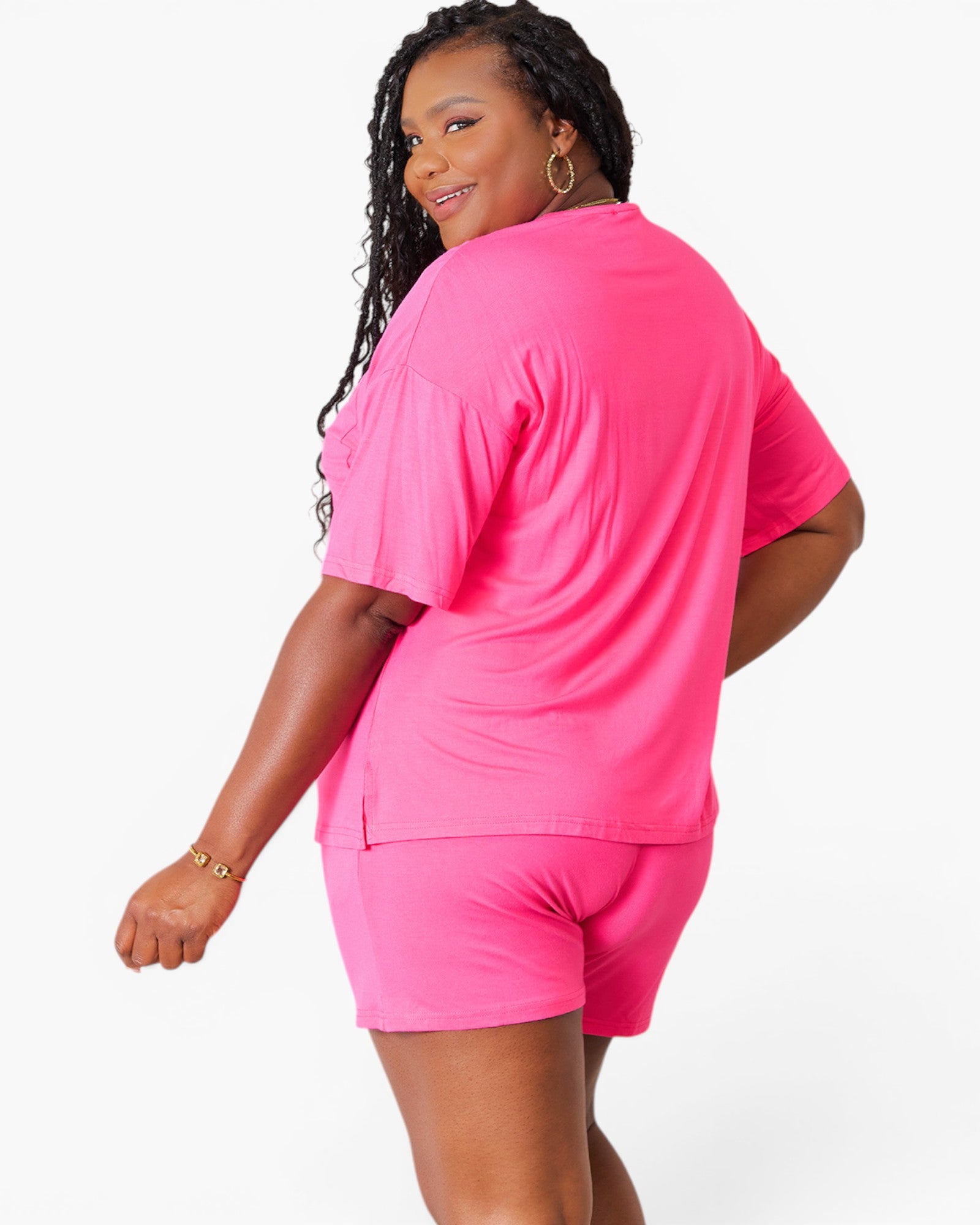 A woman modeling a bright pink short-sleeve t-shirt and matching shorts set, shown from a three-quarters back view against a plain white studio background.
