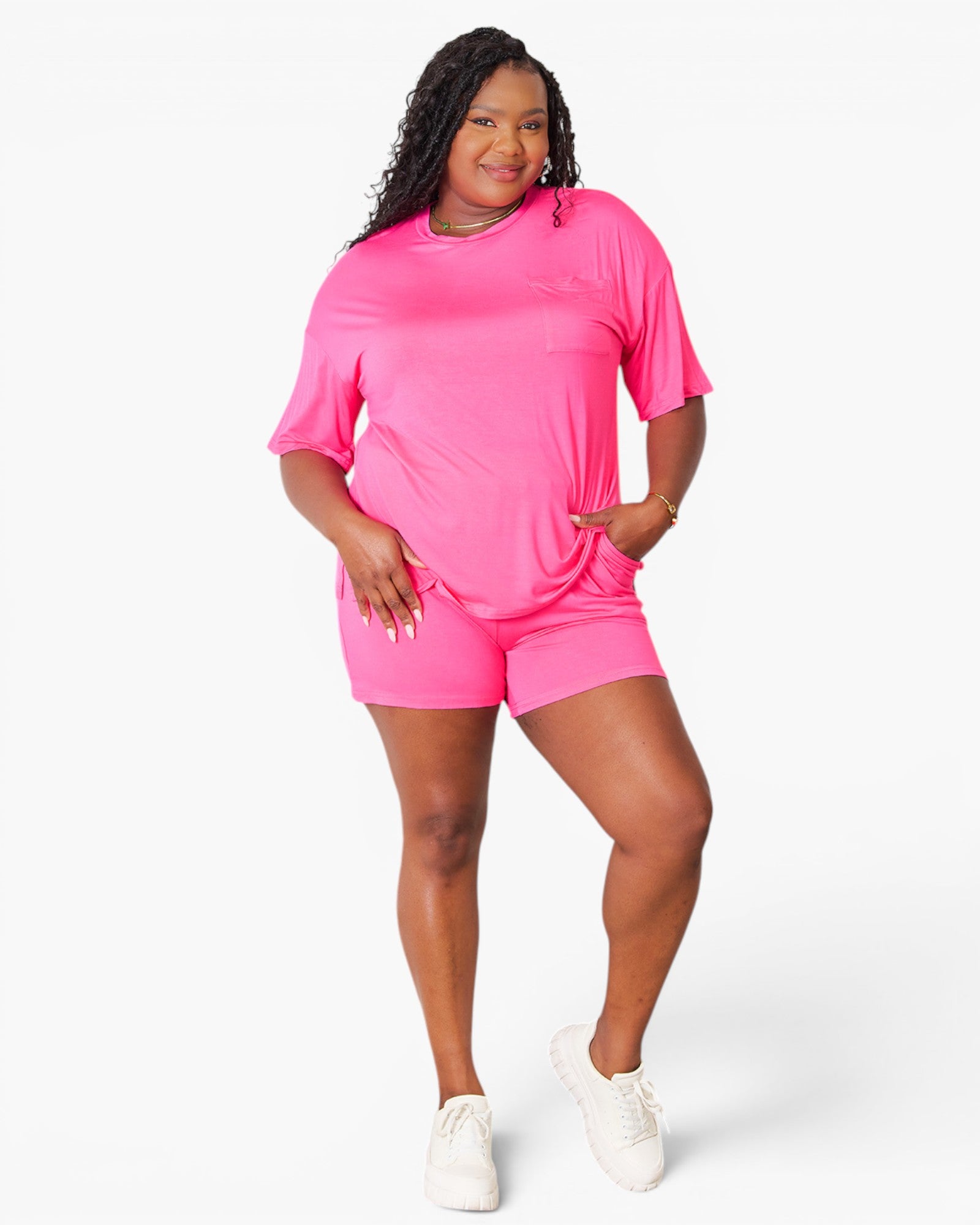 A plus-size model wearing a bright pink short-sleeve t-shirt and matching pink athletic shorts, paired with white sneakers, against a clean white studio background.