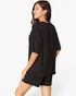 A woman wearing a black oversized short-sleeve top and matching black shorts, shown from a side-back angle against a clean white studio background.
