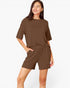 A woman modeling a matching brown lounge set consisting of a short-sleeve crewneck top and coordinating shorts, photographed against a clean white studio background.