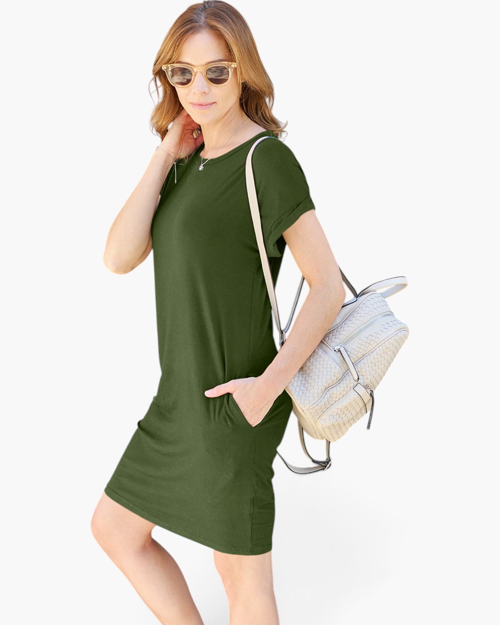 A woman modeling a short-sleeve olive green mini dress with side pockets, styled with a light-colored woven shoulder bag and sunglasses against a plain white background.