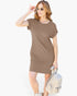 A woman modeling a short-sleeve brown t-shirt dress, holding a light beige textured handbag and wearing gold-framed sunglasses against a plain white background.