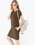 A woman modeling a short-sleeve olive brown t-shirt dress with side pockets, styled with a light-colored woven shoulder bag and sunglasses, against a plain white background.