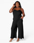 A woman modeling a black sleeveless wide-leg jumpsuit with thin spaghetti straps and side pockets, paired with clear high-heeled sandals, against a plain white studio background.