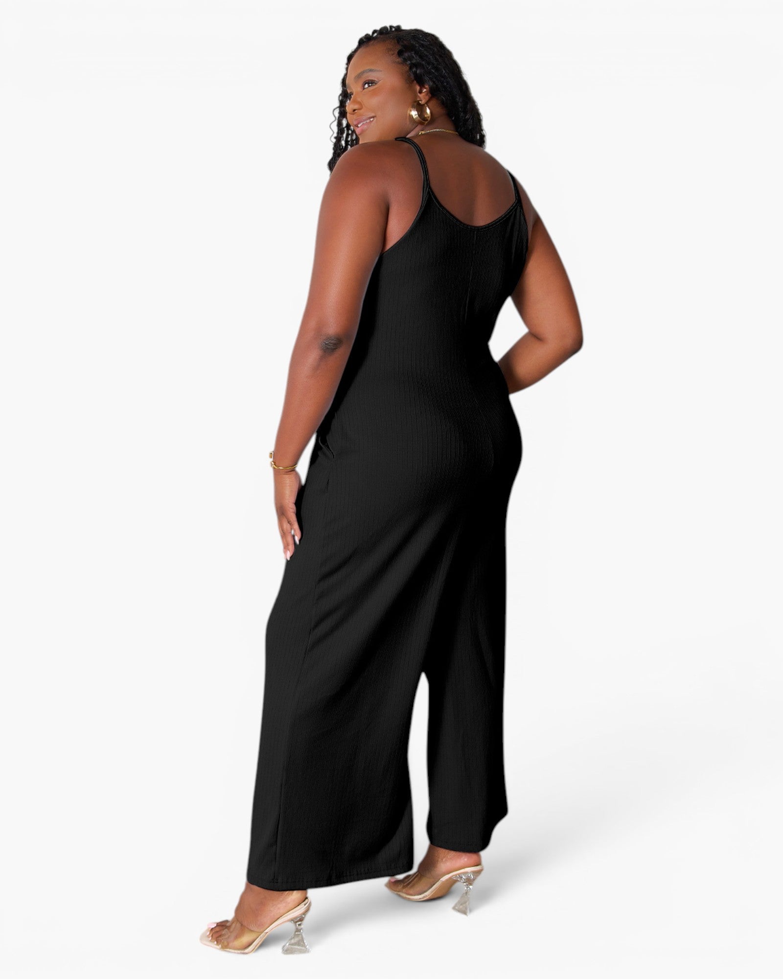 A woman modeling a black sleeveless wide-leg jumpsuit from the back, wearing clear high-heeled sandals against a plain white studio background.
