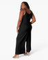 A woman modeling a black sleeveless wide-leg jumpsuit from the back, wearing clear high-heeled sandals against a plain white studio background.