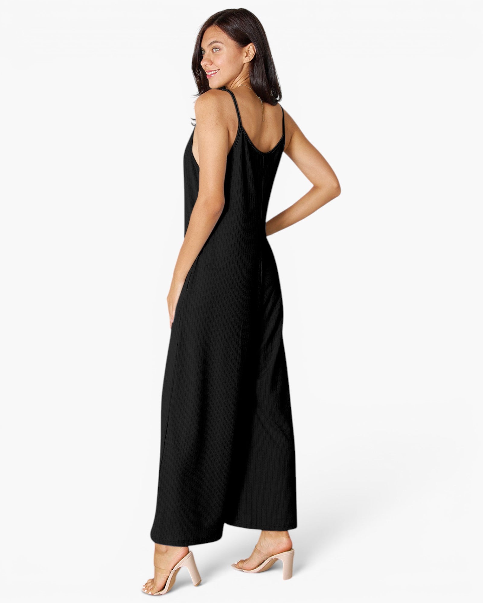 A woman modeling a black sleeveless jumpsuit from the back, wearing clear-strap high heels against a plain white studio background.