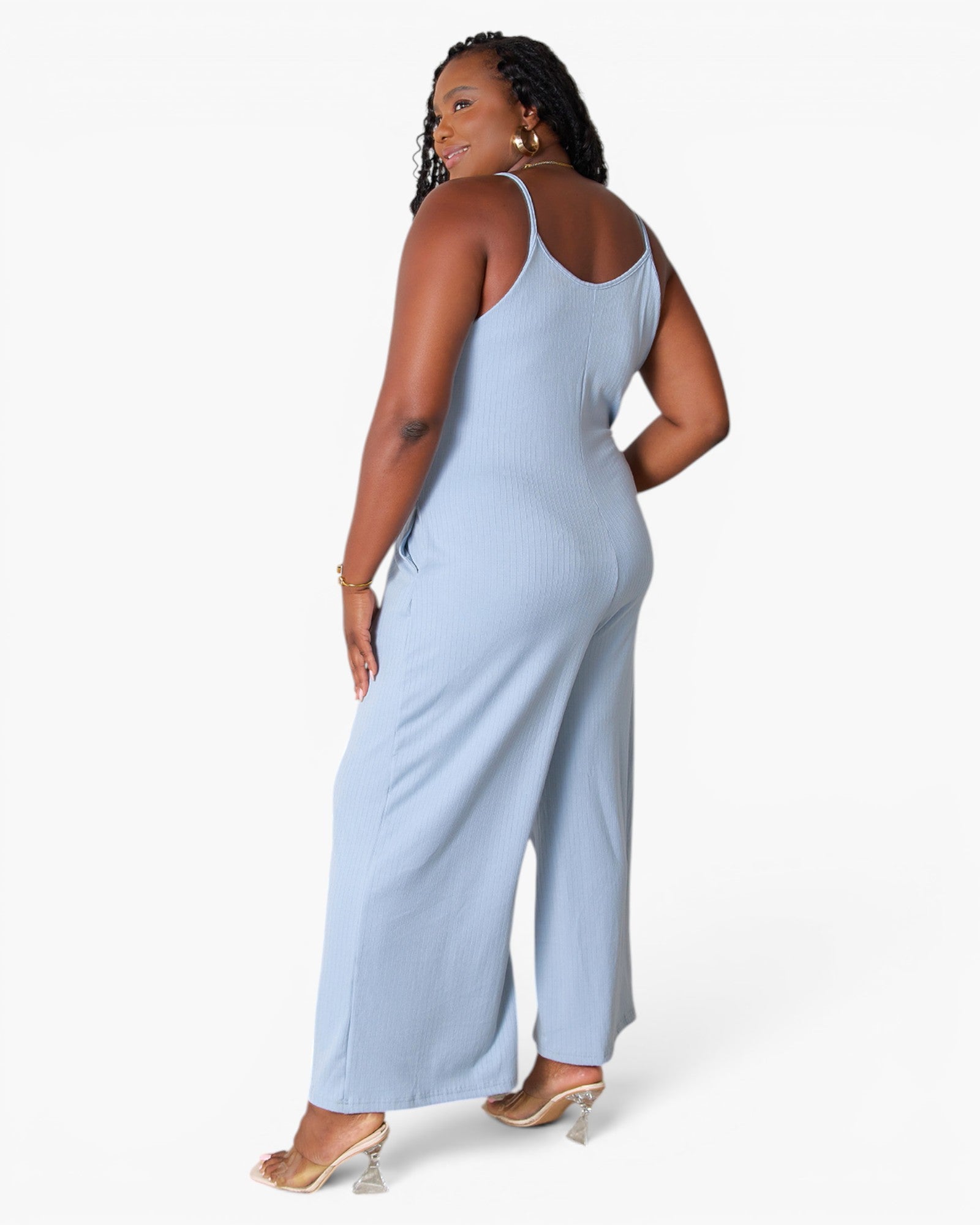 A woman modeling a light blue ribbed sleeveless jumpsuit from the back, standing against a plain white studio background.