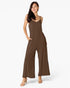 A woman modeling a sleeveless wide-leg jumpsuit in a solid dark brown color, standing against a clean white studio background.