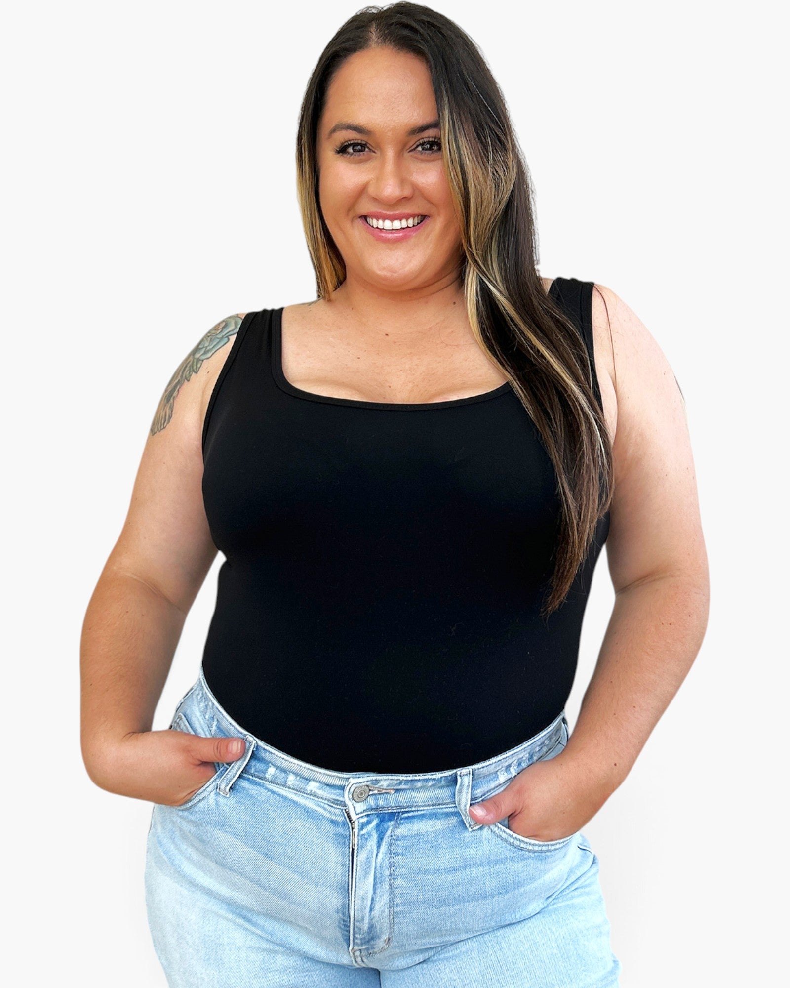 A woman wearing a black sleeveless scoop-neck tank top and light-wash blue jeans, standing against a plain white background.