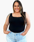A woman wearing a black sleeveless scoop-neck tank top and light-wash blue jeans, standing against a plain white background.