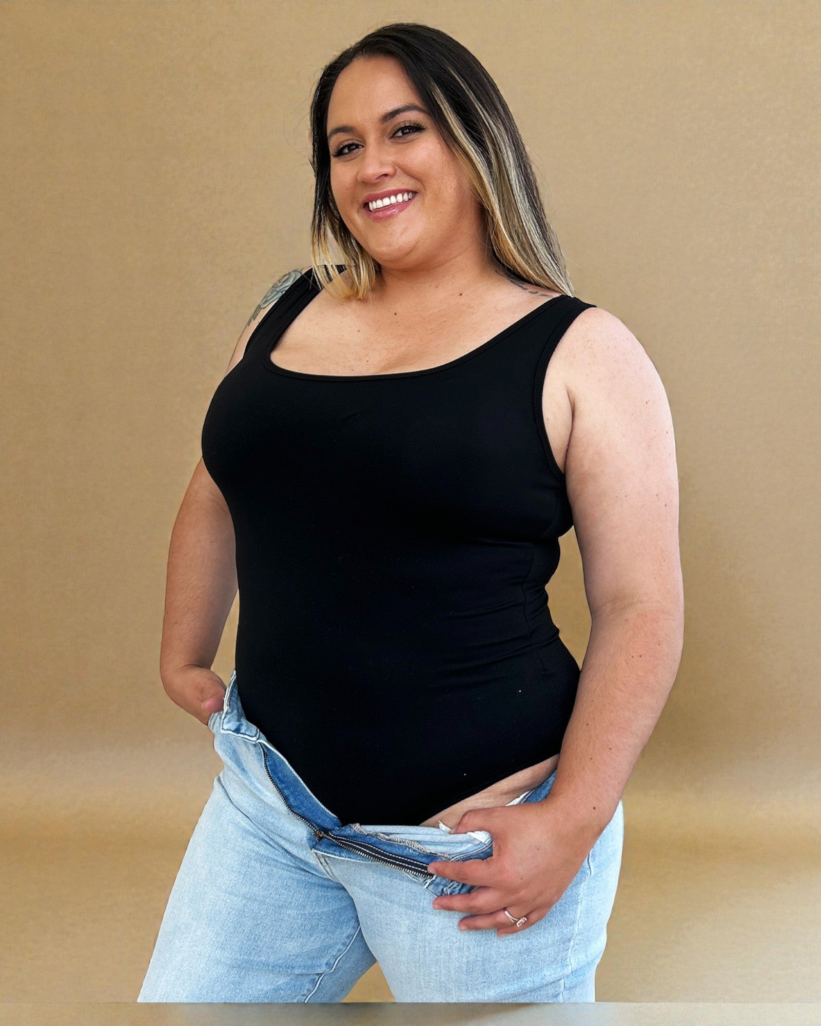 Woman wearing a black tank top and light blue jeans against a beige background